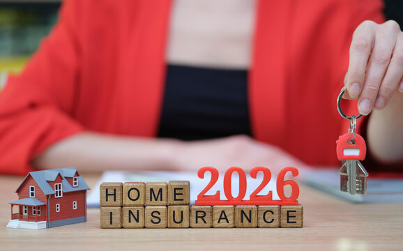In a bright office space, a person holds keys while sitting beside a cute model house and letters spelling home insurance for the year 2026. This reflects preparation for future protection.
