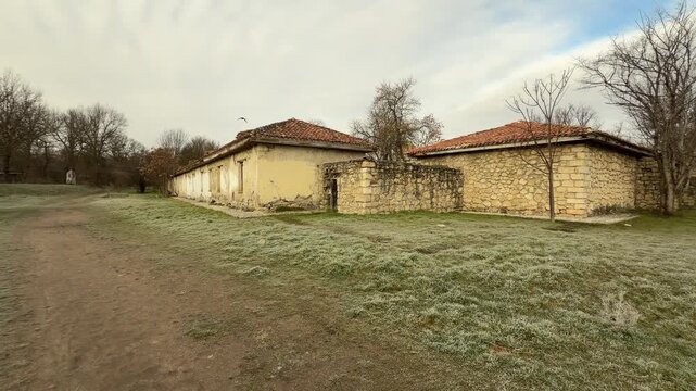 Weathered stone buildings with tiled roofs line a quiet dirt path. The surrounding grass is silver with frost, creating a lonely and nostalgic atmosphere. This winter scene feels calm and still.