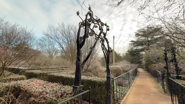 A beautiful metal sculpture shaped like a tree frames a quiet garden walkway. The surrounding bushes are covered in light frost, creating a peaceful and mysterious winter atmosphere in the park.