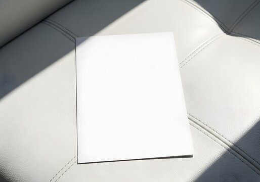 Blank white brochure or magazine mockup placed on a white tufted leather surface with a dramatic diagonal shadow cast by natural light, minimalist template for graphic design.