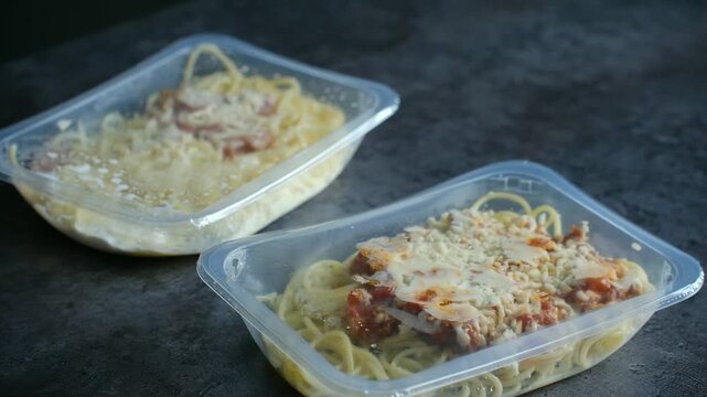 Two transparent plastic containers of takeaway spaghetti, one with bolognese sauce and one with carbonara sauce, being placed on a dark stone table by a person's hands for a quick lunch or dinner