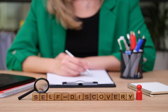 A person sits at a wooden desk, writing intently on a clipboard. Nearby, wooden blocks spell out self-discovery, reflecting a journey of personal growth amidst colorful pens and tools.