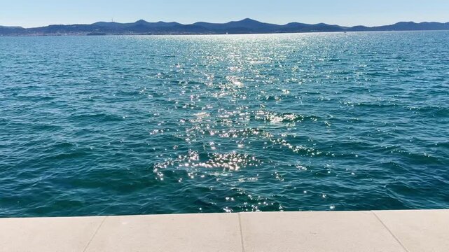 Slow‑motion shot of the shimmering turquoise Adriatic in Zadar, with a thin white stone promenade edge for contrast and distant Ugljan island on the bright horizon.
