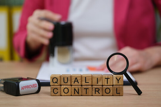 A person performs quality control checks at a desk filled with essential tools. Wooden blocks spell quality control while a magnifying glass and stamp contribute to the meticulous process.