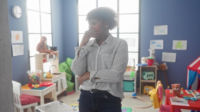Woman with arms crossed and hands covering face in kindergarten building amid toys and play tent; exhaustion patience.