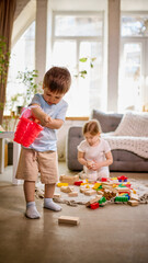 Obraz premium Little boy pouring toys from red bucket while girl builds with blocks in background. Concept of exploration, messy play, independence and natural childhood behavior.