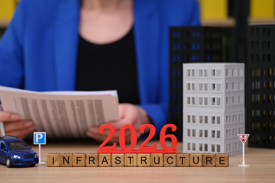 A person holds documents while focusing on a model city with blocks and symbols. Bright letters spell 2026 and infrastructure, hinting at upcoming projects and urban growth strategies.