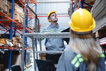 Focused worker in a yellow hard hat inspects wooden pallets on a metal ladder within a brightly lit warehouse environment while wearing a grey shirt and fluorescent green vest. © AU USAnakul