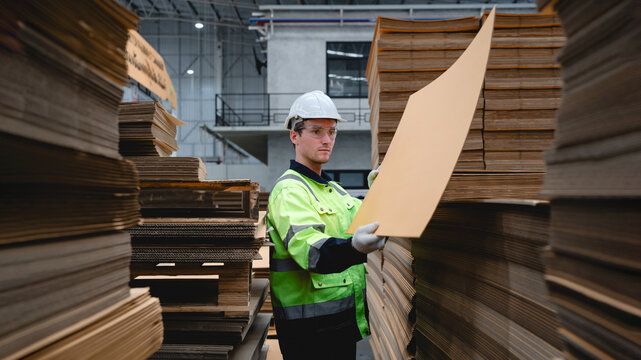 Worker in a yellow safety vest and glasses is holding up a cardboard box in a busy warehouse filled with stacked cardboard boxes, representing paper packaging and industrial logistics.