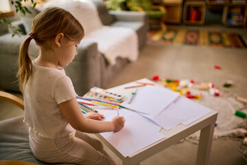 Fototapeta premium Little girl drawing with pencils at small table in cozy home interior. Concept of kids products promotion, creative classes advertising, early education materials and hobby development content.