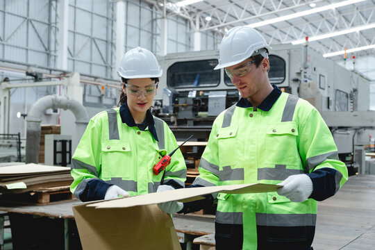 Two workers in fluorescent green high-visibility jackets and white helmets inspect a cardboard box in a bright industrial setting with machinery and a large ventilation system.