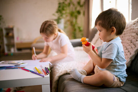 Little boy sitting on sofa holding toy while girl drawing at table during quiet playtime at home. Concept of independent play routines, parenting time management and calm home activity scenarios.