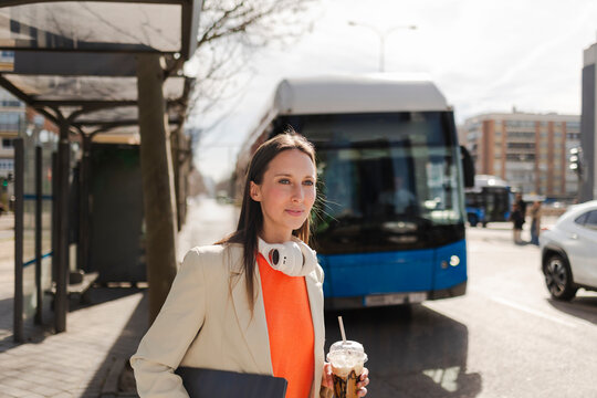 Businesswoman with headphones and iced coffee waiting at urban bus stop