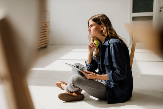 Creative woman thinking and eating apple in sunny studio with tablet