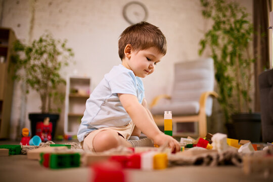 Little boy building structure with plastic blocks sitting on floor in modern living room. Concept of problem solving tasks, spatial thinking development and independent play scenarios.