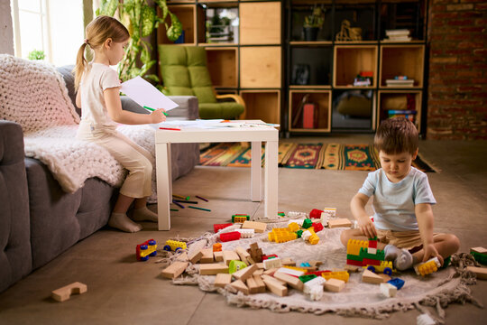 Little children playing separately at home with girl drawing and boy building blocks on floor. Concept of parallel play behavior, sibling independence and home activity routines.