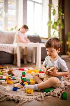 Little boy holding wooden block while sitting on floor with toys scattered around at home. Concept of toy variety usage, sensory play experience and early childhood exploration.