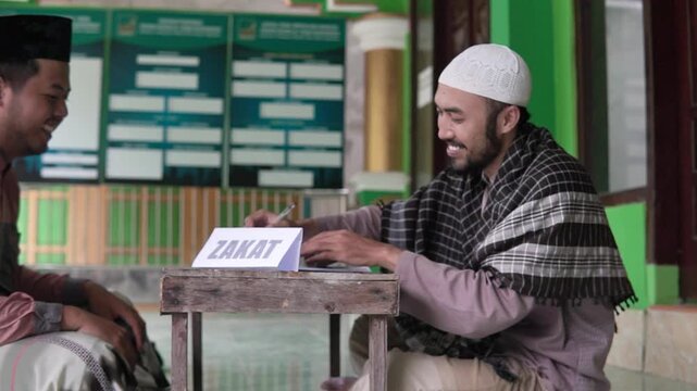 Smiling collector receives zakat donation at table, two men share handshake and laughter beside ZAKAT sign, relaxed indoor