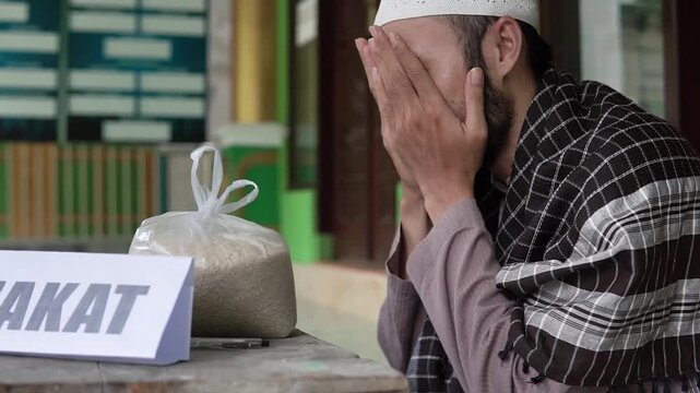 Asian man offering quiet prayer above zakat package, charity coordinator or imam tone, focused devotional posture, donation bag