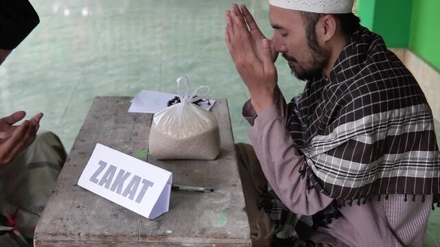 Asian man praying beside zakat rice bag, humble supplication scene, mosque veranda atmosphere, collector and donor roles