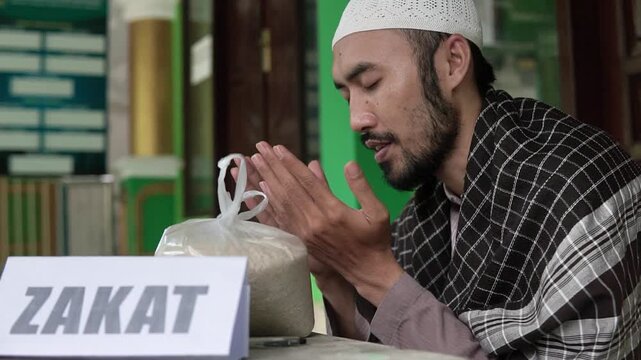 Asian muslim man praying beside zakat donation sign and rice bag on table inside mosque solemn donor portrait wearing white cap