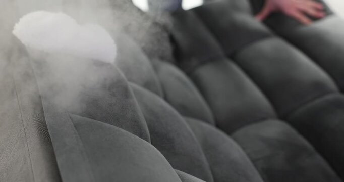 Man uses steam cleaner to carefully clean modern gray sofa. Male moves steam cleaner allowing hot vapor to refresh modern upholstery at home