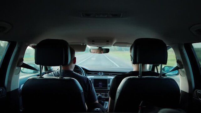 Rear view of two men inside car cabin, driver controls vehicle, passenger observes view from window, car traveling on paved road, focus on journey, road trip adventure