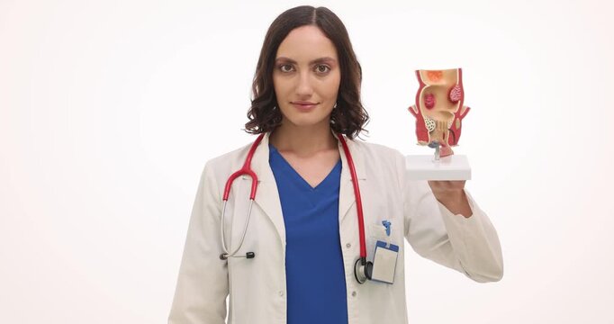 Young woman physician presents detailed model of rectum at lesson against white background. Doctor holds medical suppositories used for treatment