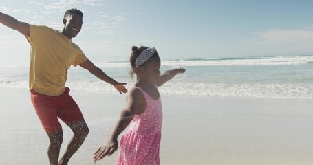 Obraz premium Running father and daughter mirroring poses, splashing surf at beach with wet sand and pink dress