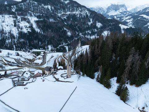 Aerial view of Cappellina di Santa Barbara and a small mountain village surrounded by snow-covered slopes and evergreen forests in Morter, Trentino-Alto Adige, Italy.
