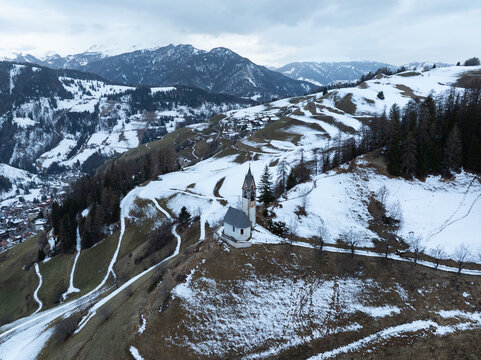 Aerial view of Cappellina di Santa Barbara on a snowy hillside with mountain peaks under an overcast sky in Morter, Trentino-Alto Adige, Italy.