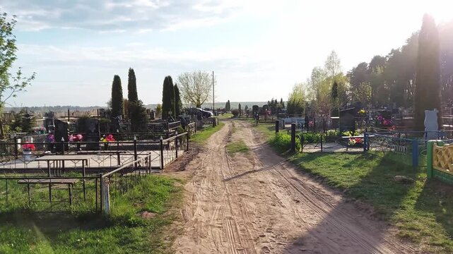 Panoramic view of peaceful burial ground featuring unpaved track winding between numerous grave sites adorned with fences and floral decorations, illuminated by warm afternoon light with serene mood