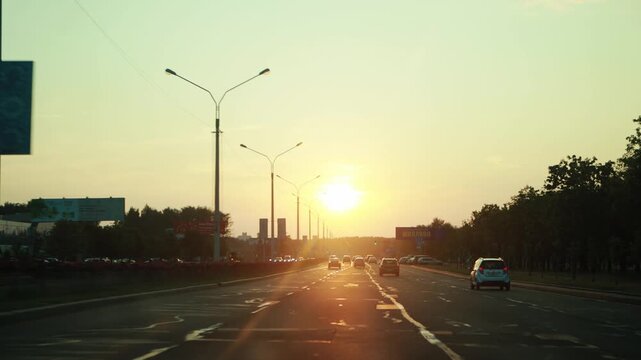 Dynamic view from moving car windshield on city street during evening. Sunset casts golden light, lens flare on asphalt road with traffic. Urban landscape includes tall buildings and streetlights.