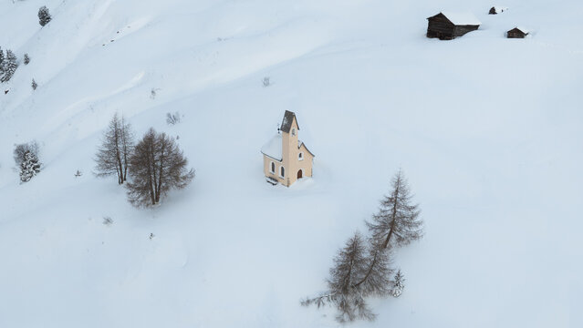 Aerial view of the Cappella di San Maurizio chapel surrounded by deep snow, pine trees, and rustic wooden barns in Selva di Val Gardena, Trentino-Alto Adige, Italy.