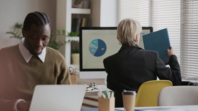 Medium shot of young Black manager working on laptop in modern office, with rear view of senior Caucasian female specialist doing paperwork in background
