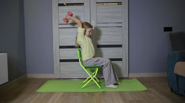Young girl with blonde hair performs overhead triceps extension with pink dumbbell. Girl seated on green chair, green yoga mat. Home fitness activity indoors.