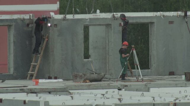 Construction workers assemble precast concrete panels. Welders join elements, sparks fly. Surveyor uses total station. Modern urban residential construction scene.