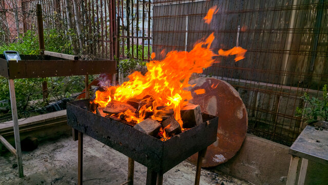 wide angle view of a square iron brazier on legs burning large wooden logs with bright orange flames in a suburban backyard yard with green plants and metal fence