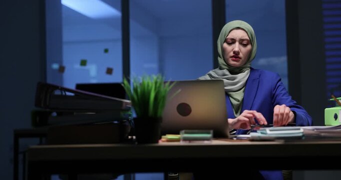 Businesswoman in hijab narrows eyes as tries to read information on laptop screen. Office worker puts on glasses to see more clearly at thunderstorm