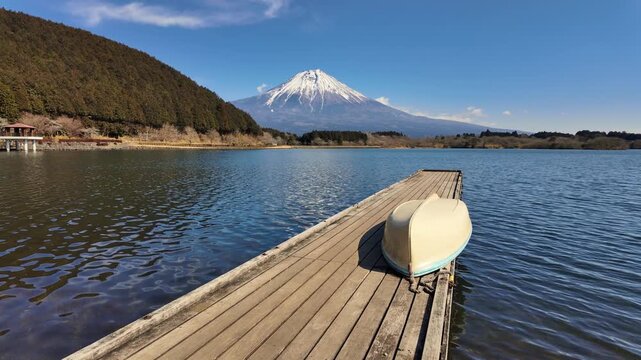 A peaceful scene at Lake Tanuki featuring a lone rowboat on a wooden jetty. The camera tilts up to reveal the magnificent snow-capped Mount Fuji under a brilliant blue sky.