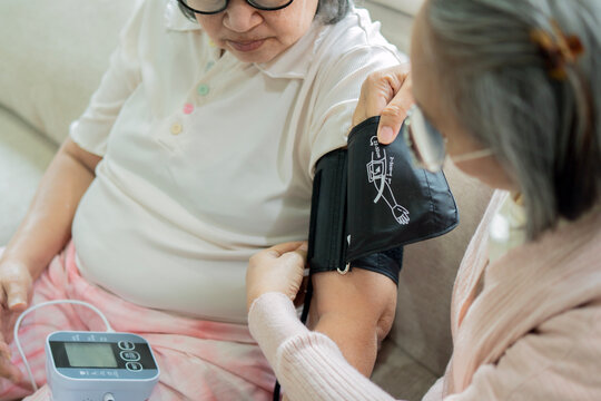 Asian female nurse measuring blood pressure of senior woman sitting on sofa at home
