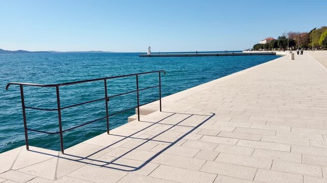 Calm slow‑sunny scene on Zadar&rsquo;s white stone promenade with distant unrecognisable people, a small white lighthouse, shimmering Adriatic water, and a black handrail casting shadows.