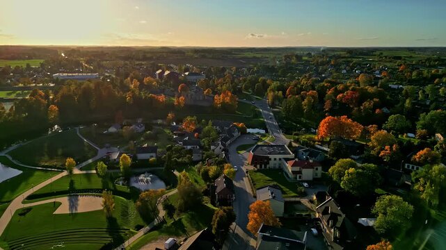 A drone flies forward over sun-drenched autumn trees and a fountain in the town of Dobele.