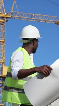 Construction Worker Reviews Site Plans. Black Engineer Inspects Plans Nearby Crane. Black Engineering Professional Carefully Analyzes Site Layout While Standing Beside Large Construction Crane