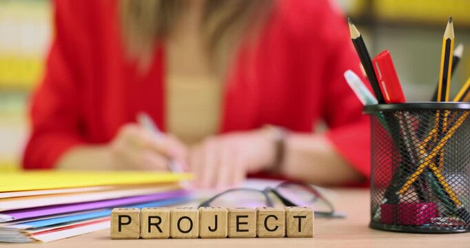 Woman office worker drafts startup project and types notes into document. Small wooden blocks on table form word Project near neat stack of papers