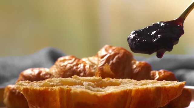 Close-up of a spoon spreading dark fruit jam onto a freshly baked croissant. Golden flaky texture and rich topping highlight a delicious breakfast moment with warm, inviting food styling.