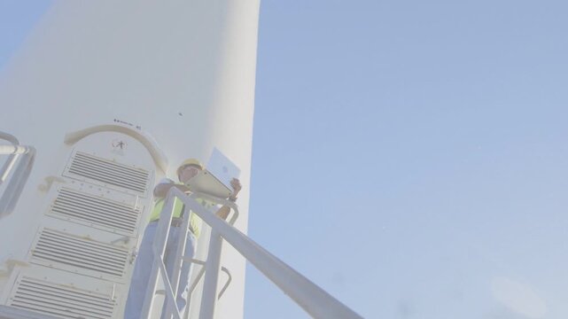 Tech checking power on laptop while camera tilting upward, live data rising wind tower to blades