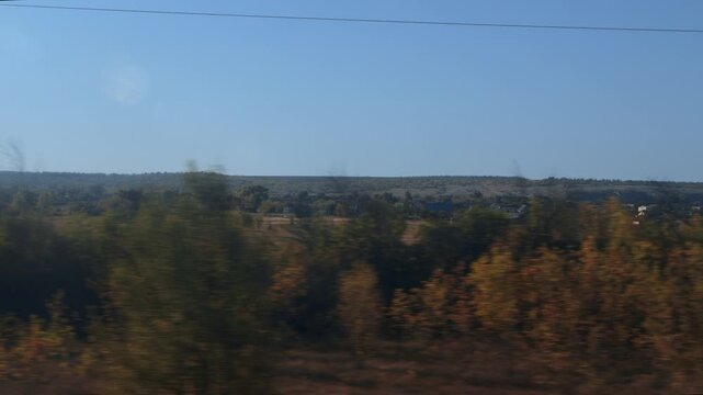 View from train window showing blurred foreground trees with golden and brown leaves. Distant small village houses visible against rolling hills. Clear blue sky above suggests bright day, journey pers