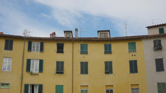 Curved row of historic yellow and white buildings in Lucca's Piazza dell'Anfiteatro, Italy. Traditional green shutters, terracotta roofs and TV antennas against a blue sky. Travel vacation in Europe