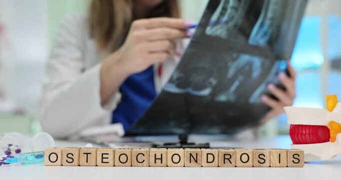 Woman physician looks closely at spine X-ray picture. Wooden cubes on table form word Osteochondrosis near anatomical model of spinal disc on table